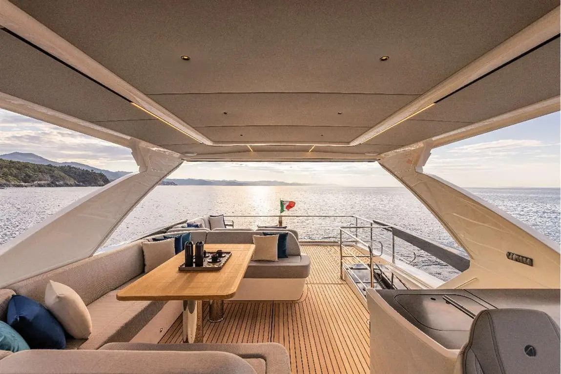 Spacious yacht deck with wooden table, soft seating, and panoramic sea views at sunset, featuring an Italian flag.